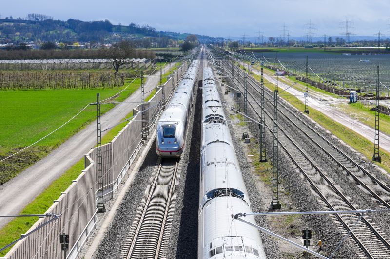 Bahngleise in einer grünen Landschaft, im Vordergrund zwei Züge, im Hintergrund ein Hügel