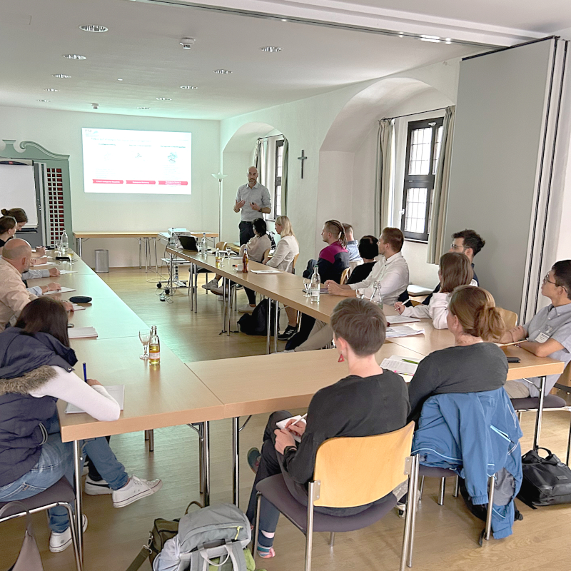 Eine Seminar-Situation: Menschen sitzen an U-förmig gestellten Tischen, vor ihnen eine Person, die einen Vortrag hält