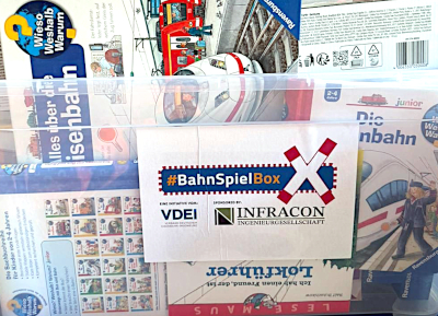 Nahaufnahme der Vorderansicht einer BahnSpielBox: eine durchsichtige Kunststoffbox, gefüllt mit Büchern und Spielzeug; vorne auf der Box die Logos des Sponsors Infracon sowie des VDEI