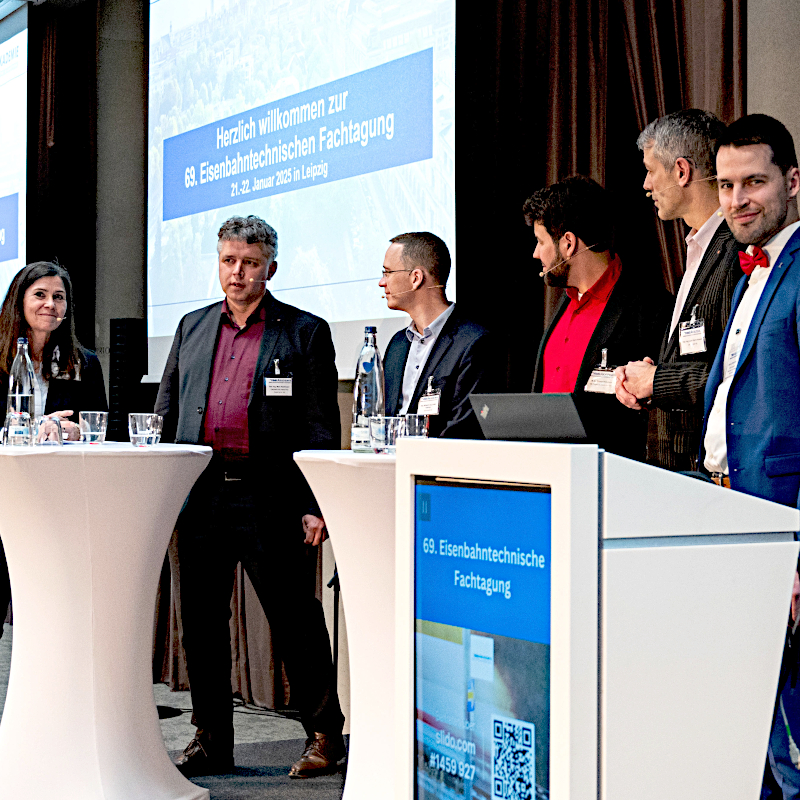 Foto von sechs Personen in Business-Kleidung, die an Stehtischen zusammenstehen.