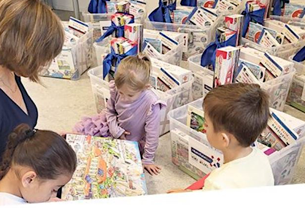 im Vordergrund drei Kinder im Kita-Alter und eine erwachsene Person, die zusammen ein Bilderbuch betrachten; im Hintergrund mehrere gefüllte BahnSpielBoxen